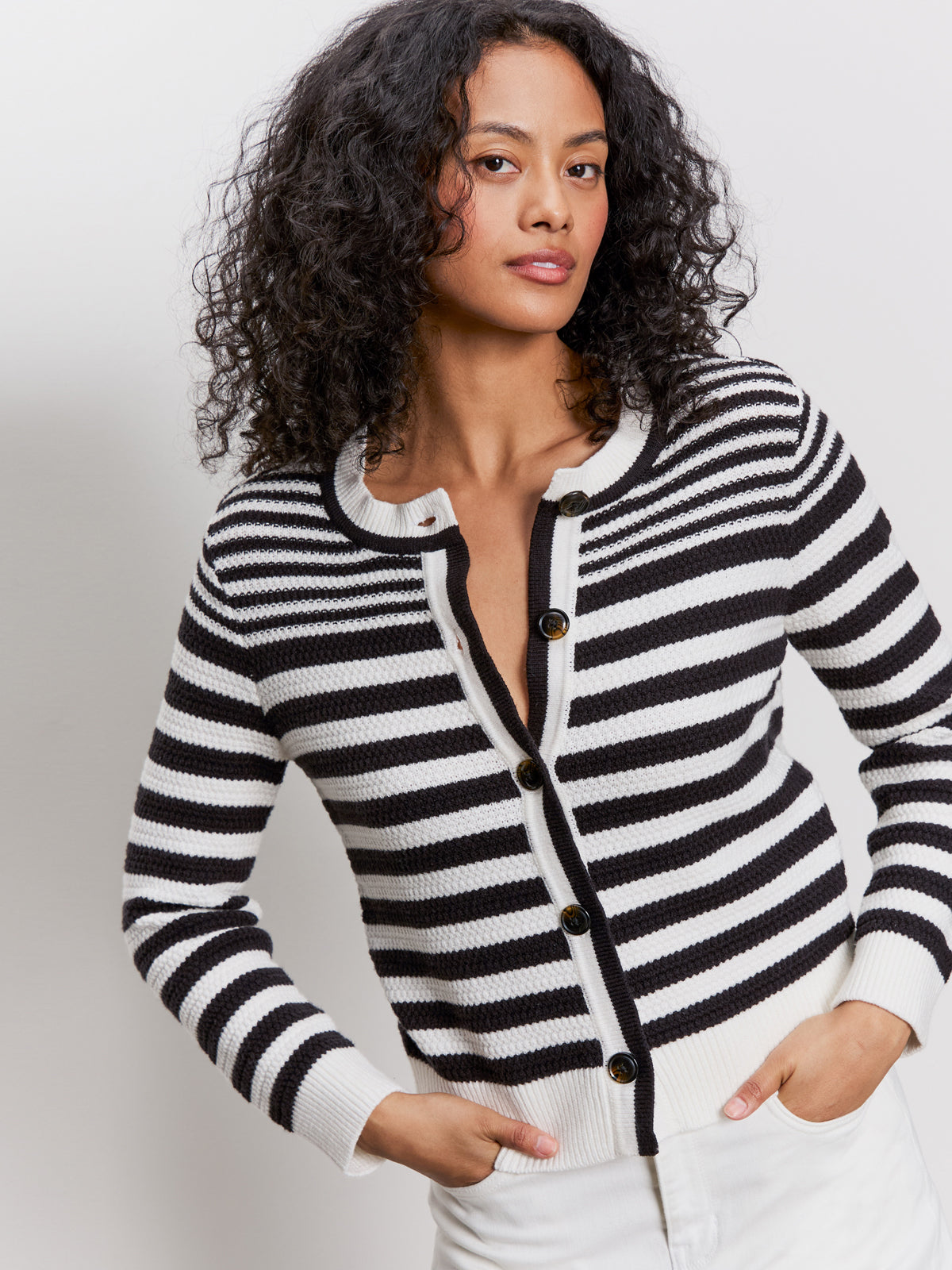 A woman with curly hair models the Sanctuary Clothing city summer cardi in black macrame multi stripe, paired with white pants, standing against a plain light background with her hands in her pockets.