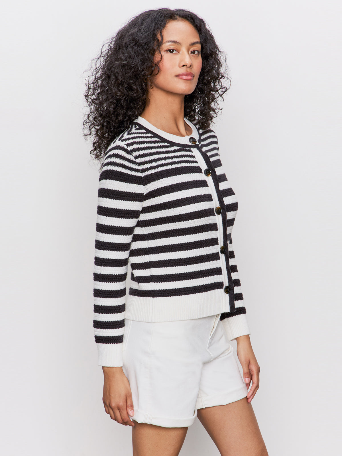 A woman with curly hair models the Sanctuary Clothing city summer cardi in black macrame multi stripe with white shorts, standing against a light background and looking at the camera.