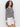 A woman with curly hair models the Sanctuary Clothing city summer cardi in black macrame multi stripe with white shorts, standing against a light background and looking at the camera.