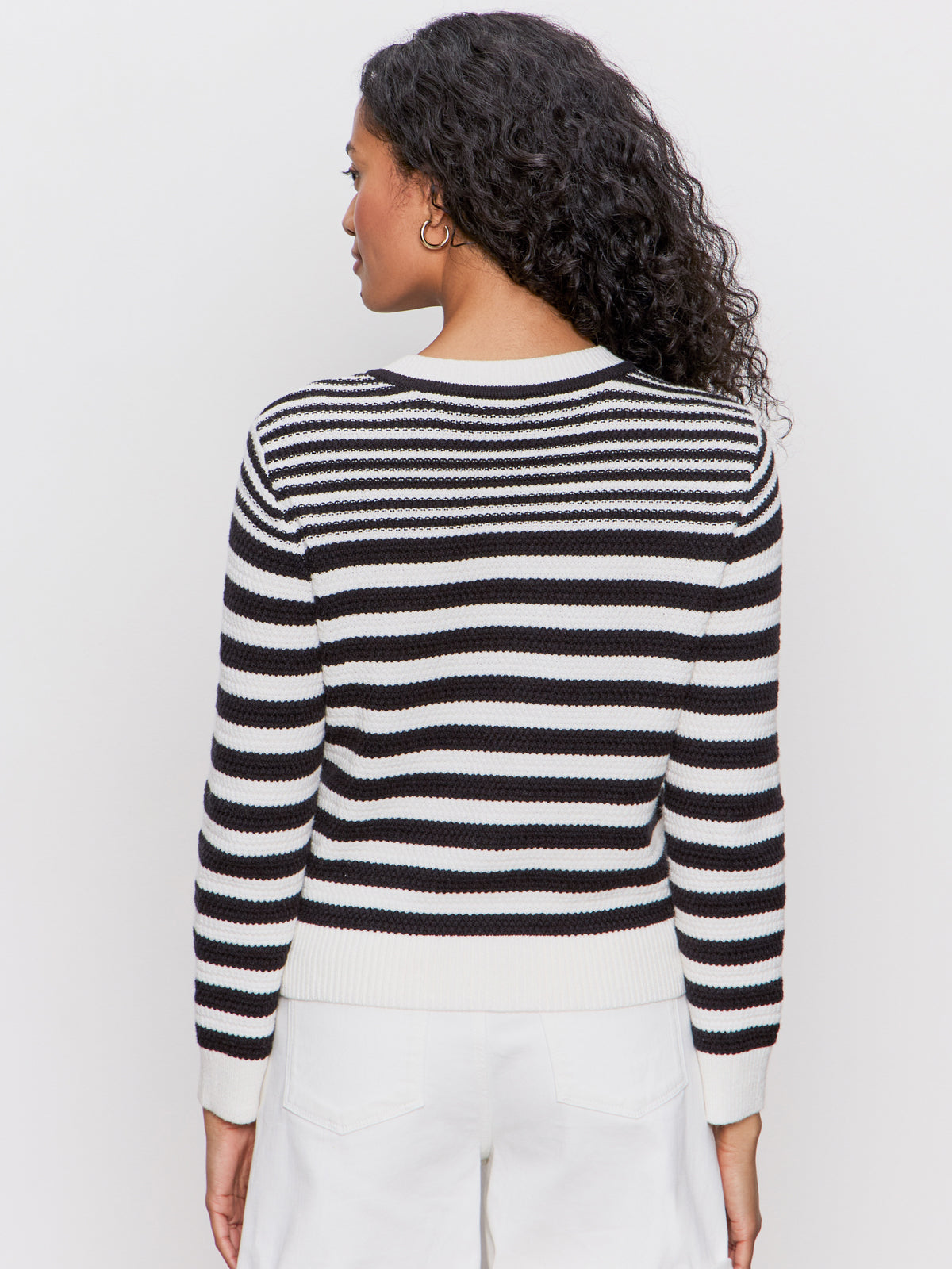 A woman with curly hair, facing away from the camera, wears white pants and the Sanctuary Clothing city summer cardi black macrame multi stripe sweater. The background is plain and light-colored.