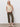 A woman with long blonde hair and a neutral expression models the Sanctuary Clothing textured sweater vest macrame against a plain white wall, styled with olive straight-leg pants and white woven slip-on shoes.