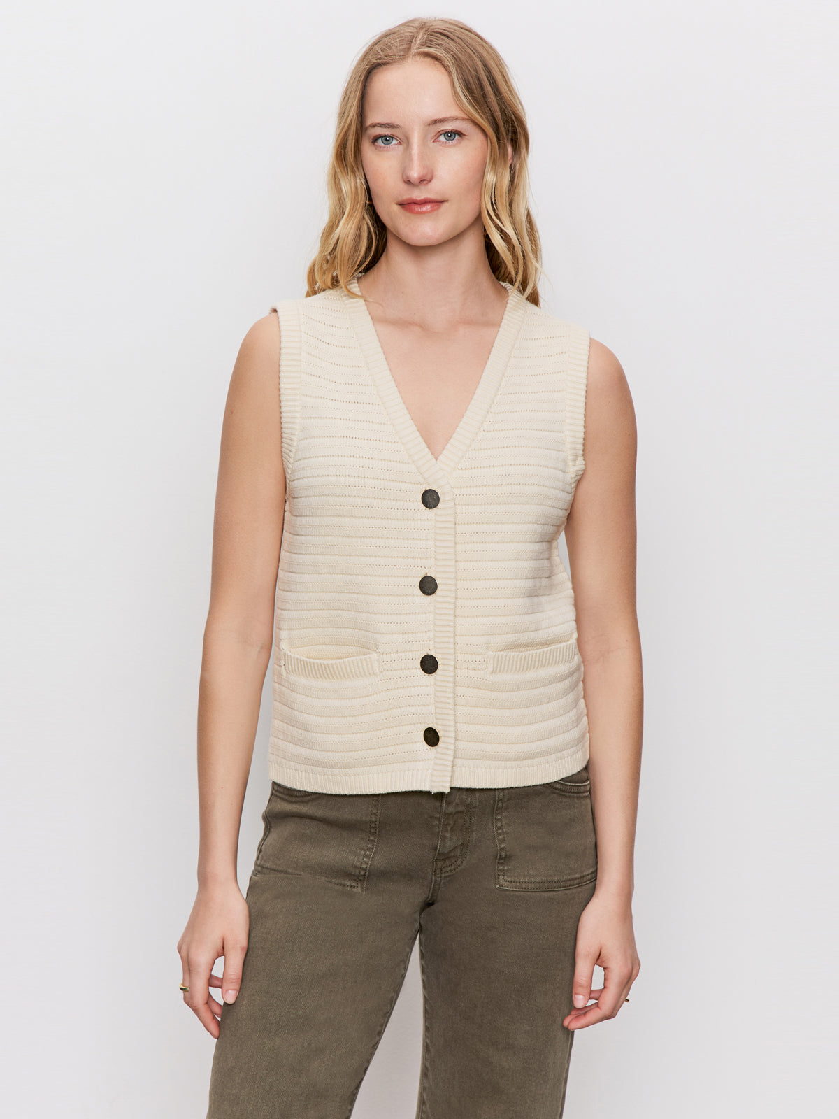 A woman with light brown hair models the Sanctuary Clothing textured sweater vest macrame—a sleeveless, cream knit design with buttons and two front pockets—paired with olive green pants against a white background.