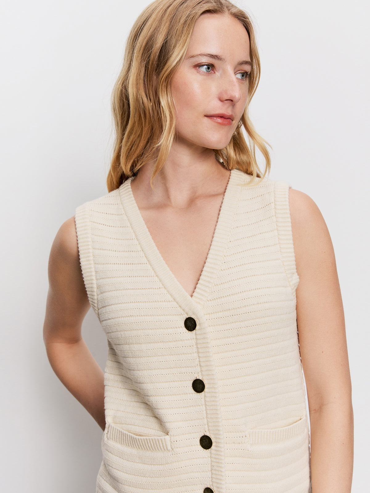 A woman with long, wavy blonde hair models the Sanctuary Clothing textured sweater vest macrame—a sleeveless, cream knit vest with black buttons and front pockets—against a plain white background, looking slightly to the side.