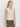 A woman with long blonde hair wears the Sanctuary Clothing textured sweater vest macrame— a sleeveless, cream-colored vest with vertical ribbing and two front pockets—paired with olive green pants against a white background.