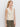 A woman with long blonde hair wears the Sanctuary Clothing textured sweater vest macrame� a sleeveless, cream-colored vest with vertical ribbing and two front pockets�paired with olive green pants against a white background.