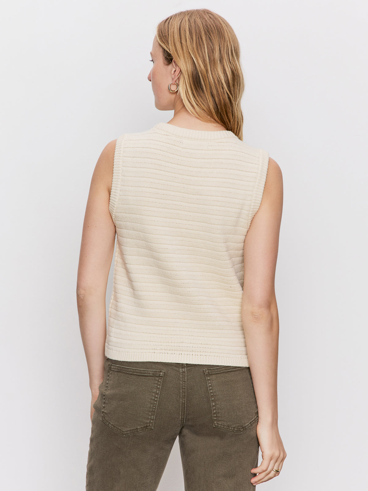 A woman with loose light brown hair stands facing away from the camera, wearing Sanctuary Clothing's textured sweater vest macrame in cream and olive green jeans against a plain white background.