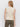 A woman with loose light brown hair stands facing away from the camera, wearing Sanctuary Clothing's textured sweater vest macrame in cream and olive green jeans against a plain white background.