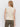 A woman with loose light brown hair stands facing away from the camera, wearing Sanctuary Clothing's textured sweater vest macrame in cream and olive green jeans against a plain white background.