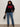A woman with curly hair wears the Sanctuary Clothing après ski sweater in black mars red (extended sizing), paired with blue jeans, black boots, and a black handbag as she stands against a plain light-colored wall.