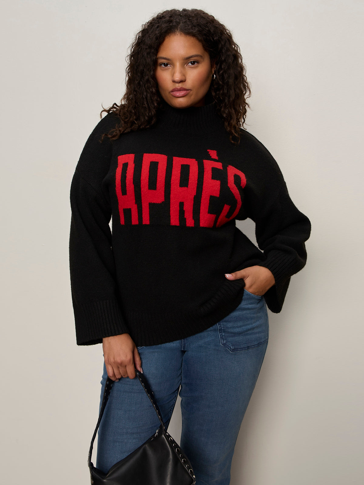 A woman with curly hair models the Sanctuary Clothing après ski sweater in black mars red (extended sizing) with bold "APRÈS" text, styled with blue jeans and a black handbag against a plain wall, looking confidently at the camera.