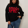 A woman with curly hair models the Sanctuary Clothing après ski sweater in black mars red (extended sizing) with bold "APRÈS" text, styled with blue jeans and a black handbag against a plain wall, looking confidently at the camera.