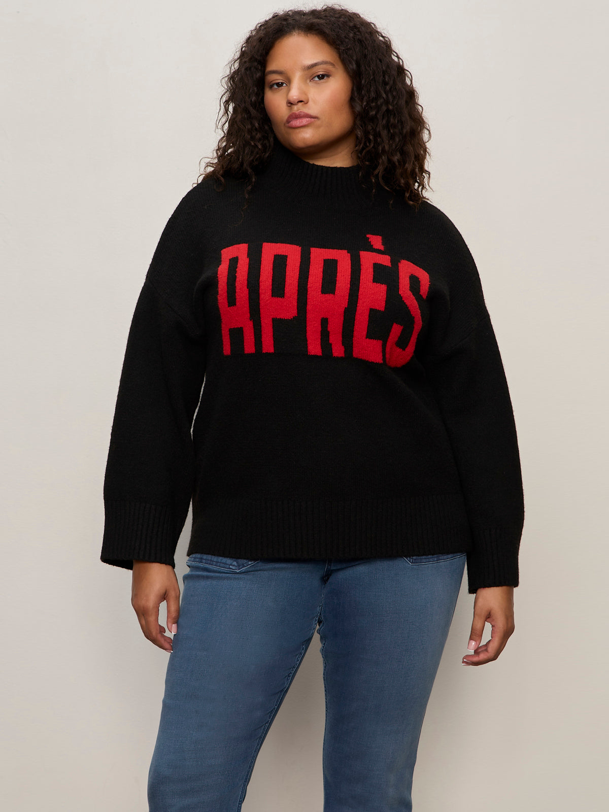 A person with curly hair wears Sanctuary Clothing's apres ski sweater in black mars red (extended sizing), featuring bold "APRÈS" lettering, styled with blue jeans against a plain light background.