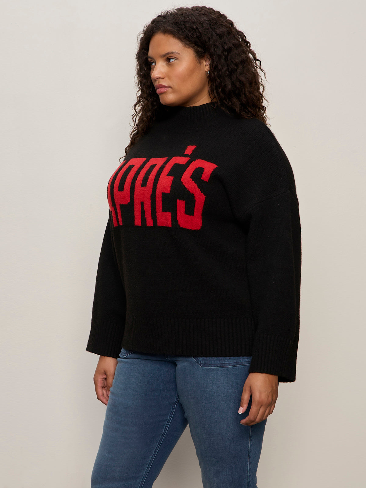 A woman with curly hair wears the Sanctuary Clothing "apres ski sweater" in black mars red (extended sizing), featuring bold red "APRÈS" lettering, paired with blue jeans against a plain, light background.