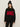 A woman with long brown hair models the Sanctuary Clothing "apres intarsia high mock logo sweater" in black/mars red, featuring bold red "APRÈS" lettering, paired with a black knit pom-pom hat and black pants against a light background.