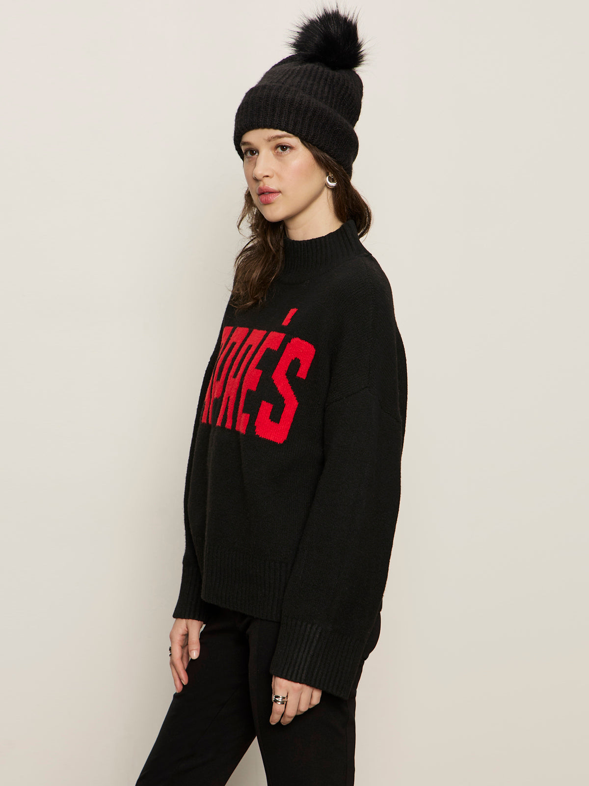 A woman models the Sanctuary Clothing apres ski sweater in black mars red (petite sizing) with a knit beanie and black pants, standing against a light background and looking at the camera.