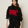 A woman stands against a plain light background wearing the Sanctuary Clothing Après Ski Sweater in black/mars red (petite sizing) and a black knit pom-pom beanie, looking off to the side in a relaxed pose.