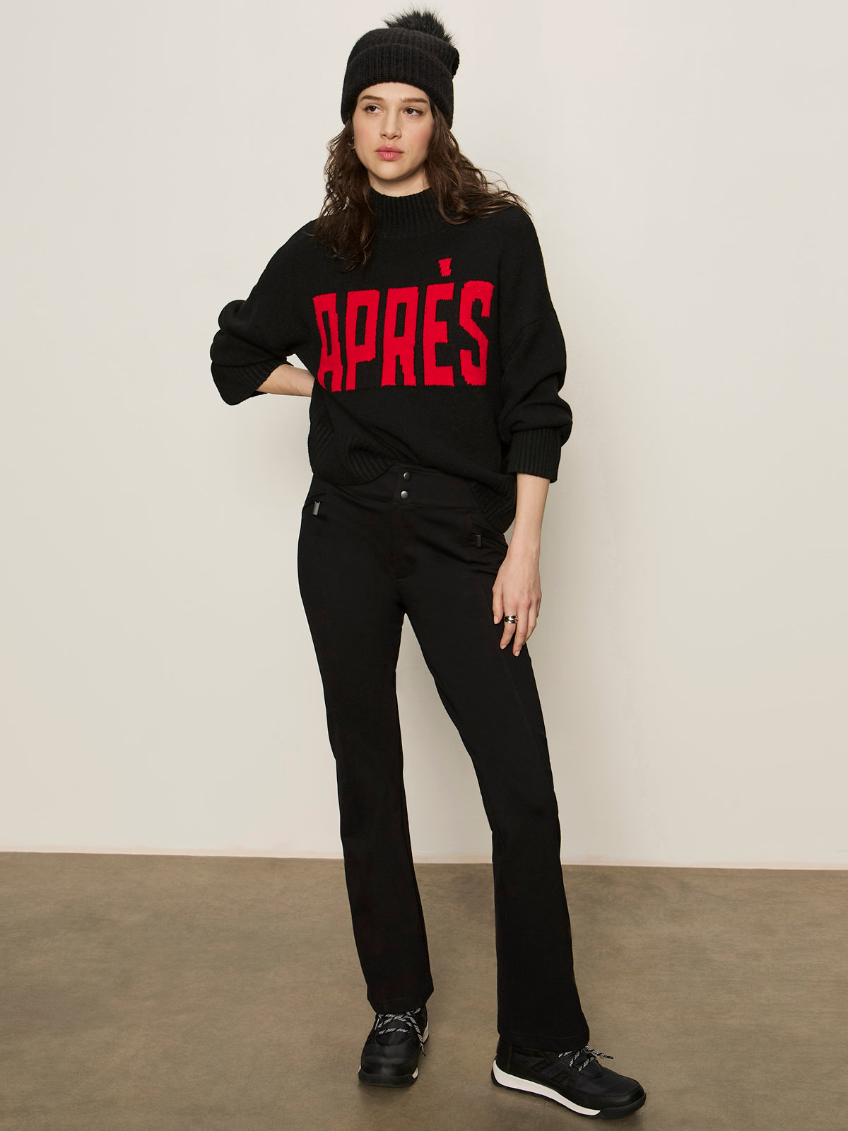 A woman stands against a plain background wearing the Sanctuary Clothing apres intarsia high mock logo sweater in black/mars red, paired with a black knit beanie, black pants, and sneakers. She has a neutral expression and one hand in her pocket.