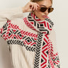 A woman poses against a plain background wearing the Sanctuary Clothing send it scarf in chalk multi, styled with aviator sunglasses and a cream knit sweater featuring red and black geometric patterns.