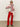 A person wears the Sanctuary Clothing send it scarf chalk multi, sunglasses, a white sweater with red and black geometric patterns, red pants, and black boots, standing against a plain background with one hand on their hip.