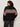 A person with curly hair stands facing away from the camera against a light background, wearing Sanctuary Clothing's send it sweater black multi extended sizing, featuring red, white, and gray geometric patterns, with dark pants.