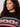 A person with long curly hair wears the "send it sweater black multi extended sizing" by Sanctuary Clothing, featuring a red and white geometric chest pattern. They look off to the side with a neutral expression.