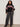 A woman wears the Sanctuary Clothing send it sweater black multi extended sizing with colorful geometric patterns, paired with dark wide-leg jeans and black ankle boots, standing against a plain light background.