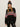 A woman wears the Sanctuary Clothing "send it sweater black multi" with white and red patterns, paired with black leather pants and a black beanie with a brown pom-pom, standing against a plain light background.