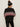 A woman stands facing away from the camera, wearing the Sanctuary Clothing send it sweater black multi and black leather pants, paired with a black winter hat featuring a white pattern and brown pom-pom.