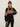 A woman models the Sanctuary Clothing "send it sweater black multi"€�a black knit with red, white, and pink geometric patterns€�and a matching beanie, posing confidently against a plain background.