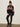A woman wearing the Sanctuary Clothing send it sweater black multi stands against a plain light-colored wall, styled with shiny black leggings, winter boots, and a black pom-pom beanie.