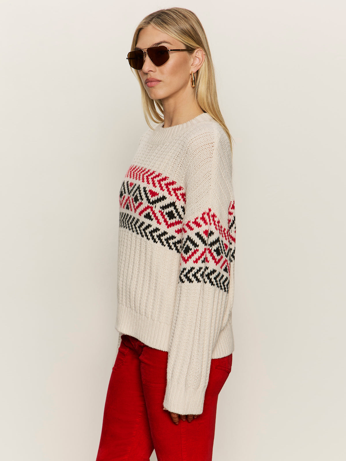 A woman wearing Sanctuary Clothing's Send It Sweater in Chalk Multi, featuring a red and black geometric pattern, along with red pants and sunglasses, stands against a plain light background.