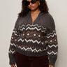 A woman with curly hair wears sunglasses, gold hoop earrings, and the Sanctuary Clothing zip up fairisle hoodie mocha fairisle extended sizing, styled with dark pants against a plain light background.