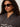 A woman with curly hair wears large sunglasses, gold hoop earrings, and the Sanctuary Clothing zip up fairisle hoodie mocha fairisle extended sizing as she looks confidently to the side against a neutral background.