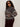 A woman with curly hair and sunglasses stands against a light background in dark pants and the Sanctuary Clothing zip up fairisle hoodie mocha fairisle extended sizing, featuring a gray and brown patterned knit, with her hand in her pocket.