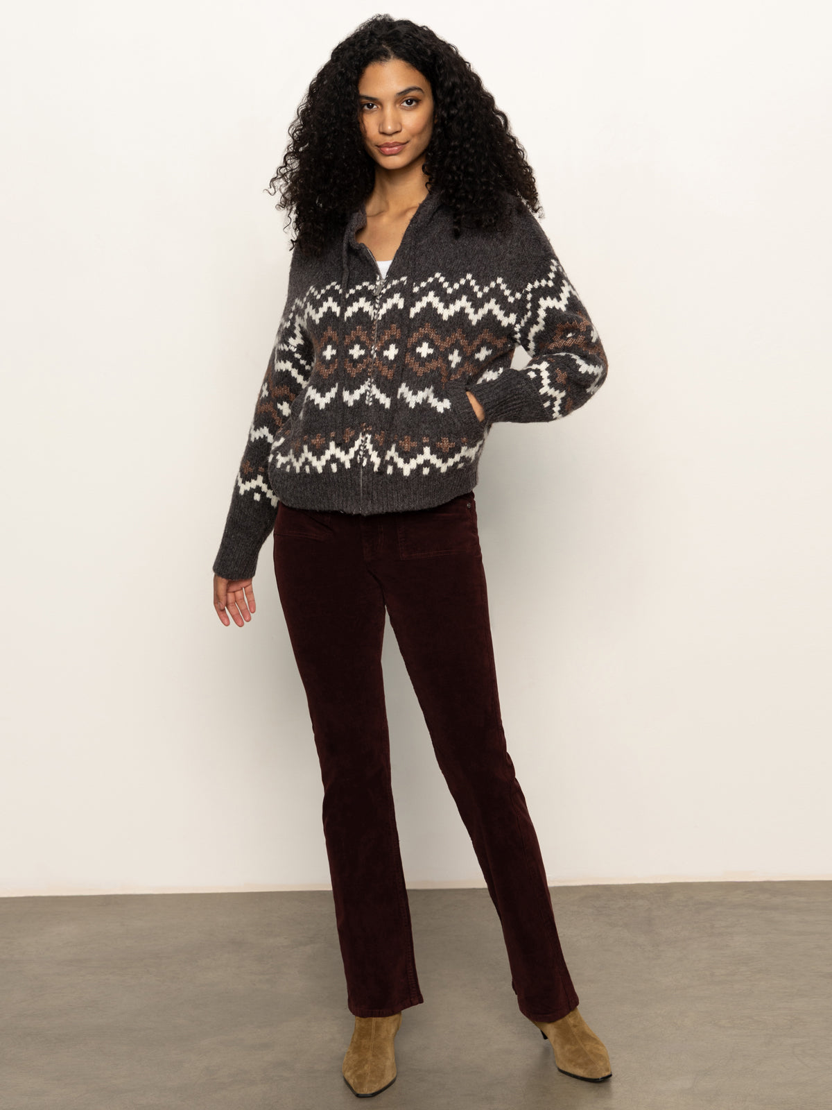A woman with curly hair wears the Sanctuary Clothing Zip-Up Fairisle Hoodie in Mocha Fairisle, paired with burgundy pants and tan shoes, standing against a light background with one hand in her pocket, looking at the camera.