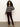 A woman with curly hair wears the Sanctuary Clothing Zip-Up Fairisle Hoodie in Mocha Fairisle, paired with burgundy pants and tan shoes, standing against a light background with one hand in her pocket, looking at the camera.