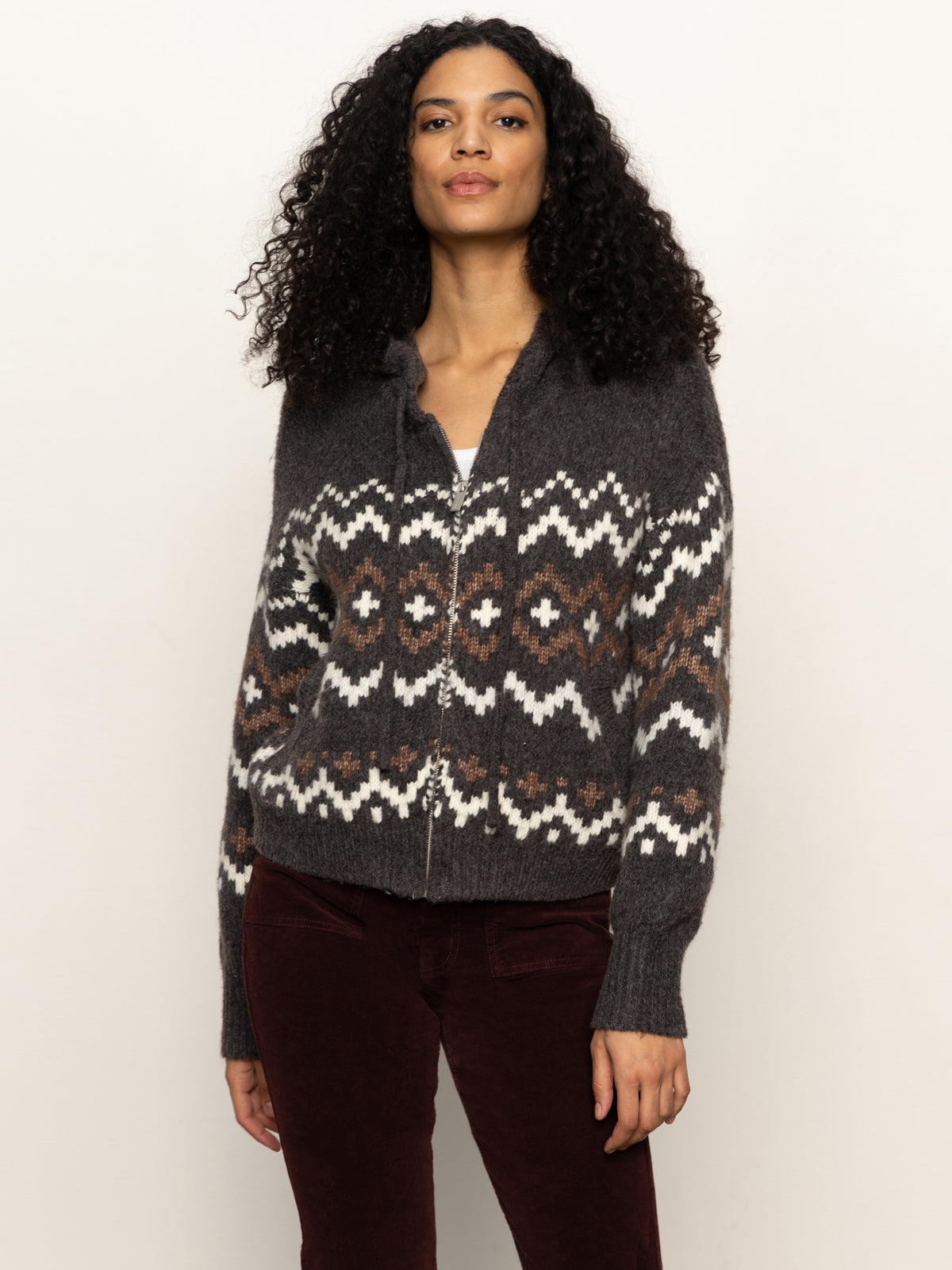 A woman with curly hair wears the Sanctuary Clothing Zip-Up Fairisle Hoodie in Mocha Fairisle over a white top and dark pants, standing against a plain light background.