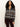 A woman with curly hair wears the Sanctuary Clothing Zip-Up Fairisle Hoodie in Mocha Fairisle over a white top and dark pants, standing against a plain light background.