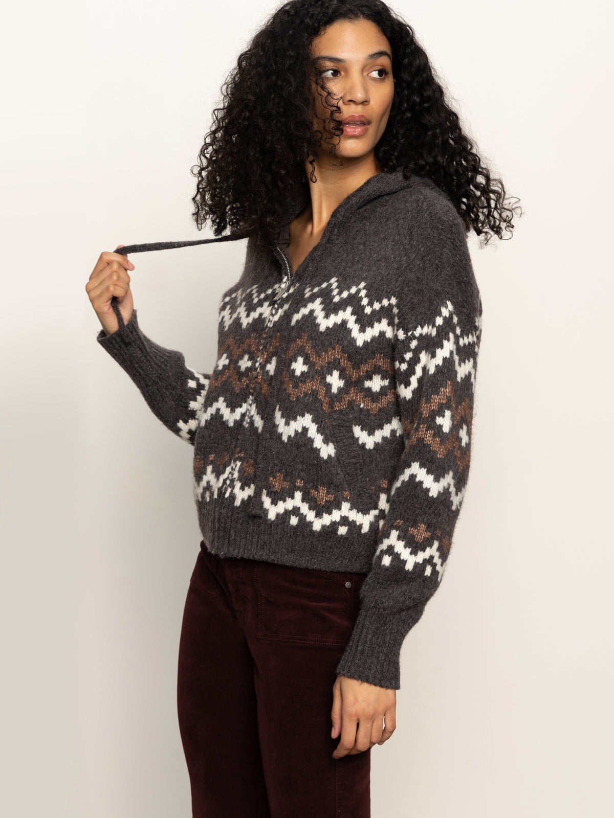 A woman with curly hair wears the Sanctuary Clothing Zip-Up Fairisle Hoodie Mocha Fairisle, featuring a white and brown geometric pattern. She holds one of the hoodie’s strings and looks to her left, paired with dark pants.