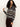 A woman with curly hair wears the Sanctuary Clothing Zip-Up Fairisle Hoodie Mocha Fairisle, featuring a white and brown geometric pattern. She holds one of the hoodie’s strings and looks to her left, paired with dark pants.