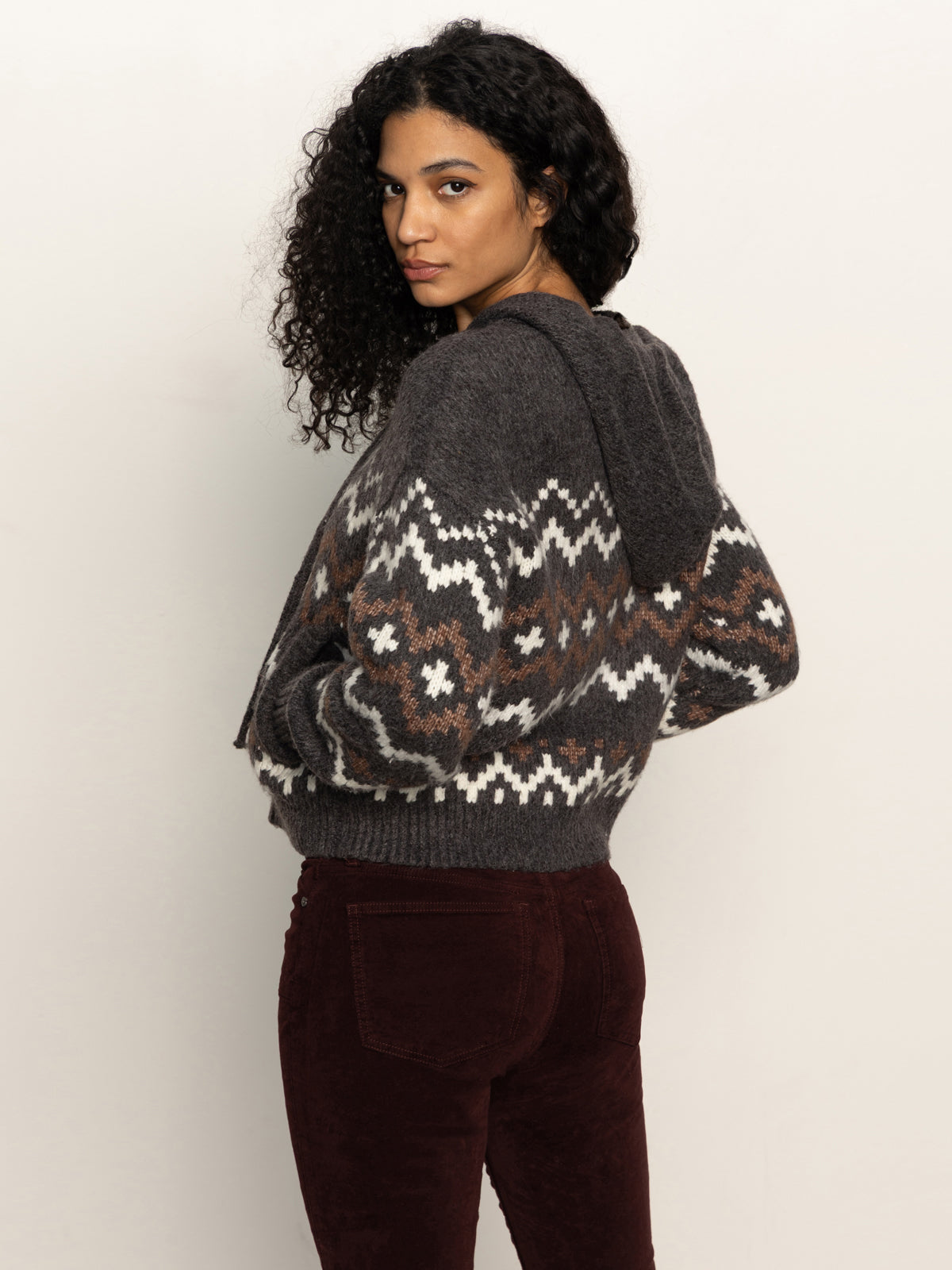 A woman with curly hair wears the Sanctuary Clothing Zip-Up Fairisle Hoodie in Mocha Fairisle, styled with dark red pants, standing sideways and looking over her shoulder against a plain background.