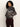 A woman with curly hair wears the Sanctuary Clothing Zip-Up Fairisle Hoodie in Mocha Fairisle, styled with dark red pants, standing sideways and looking over her shoulder against a plain background.