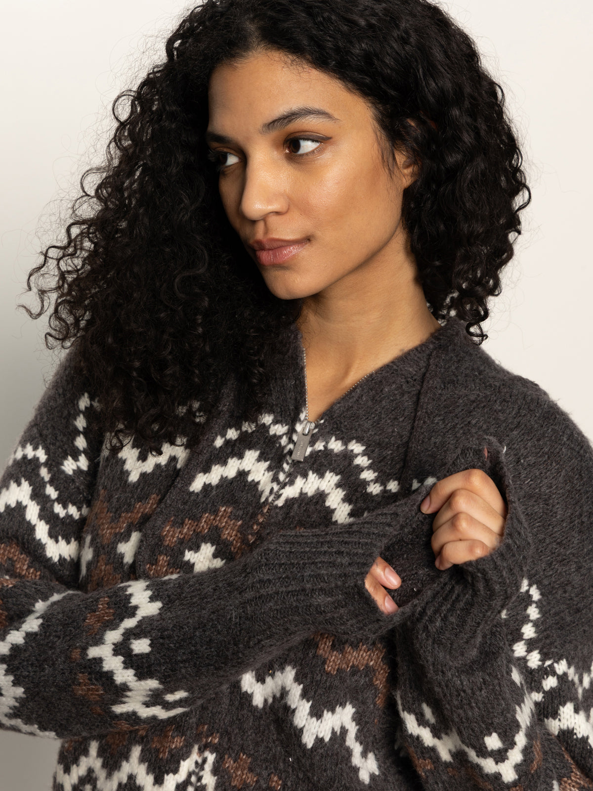 A woman with curly dark hair wears the Sanctuary Clothing Zip-Up Fairisle Hoodie in Mocha Fairisle, featuring a white and brown geometric pattern, as she looks thoughtfully to the side holding her sleeve.