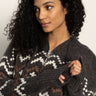 A woman with curly dark hair wears the Sanctuary Clothing Zip-Up Fairisle Hoodie in Mocha Fairisle, featuring a white and brown geometric pattern, as she looks thoughtfully to the side holding her sleeve.