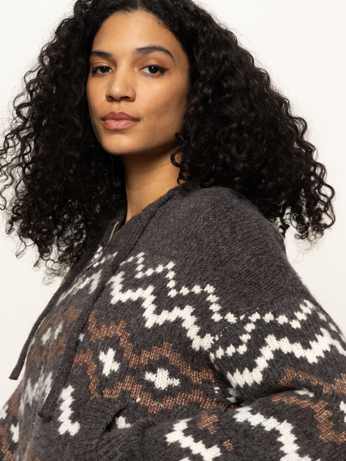 A woman with long, curly dark hair models the Sanctuary Clothing Zip-Up Fairisle Hoodie in Mocha Fairisle, featuring white and brown geometric patterns, as she looks confidently at the camera against a plain light background.