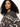 A woman with long, curly dark hair models the Sanctuary Clothing Zip-Up Fairisle Hoodie in Mocha Fairisle, featuring white and brown geometric patterns, as she looks confidently at the camera against a plain light background.