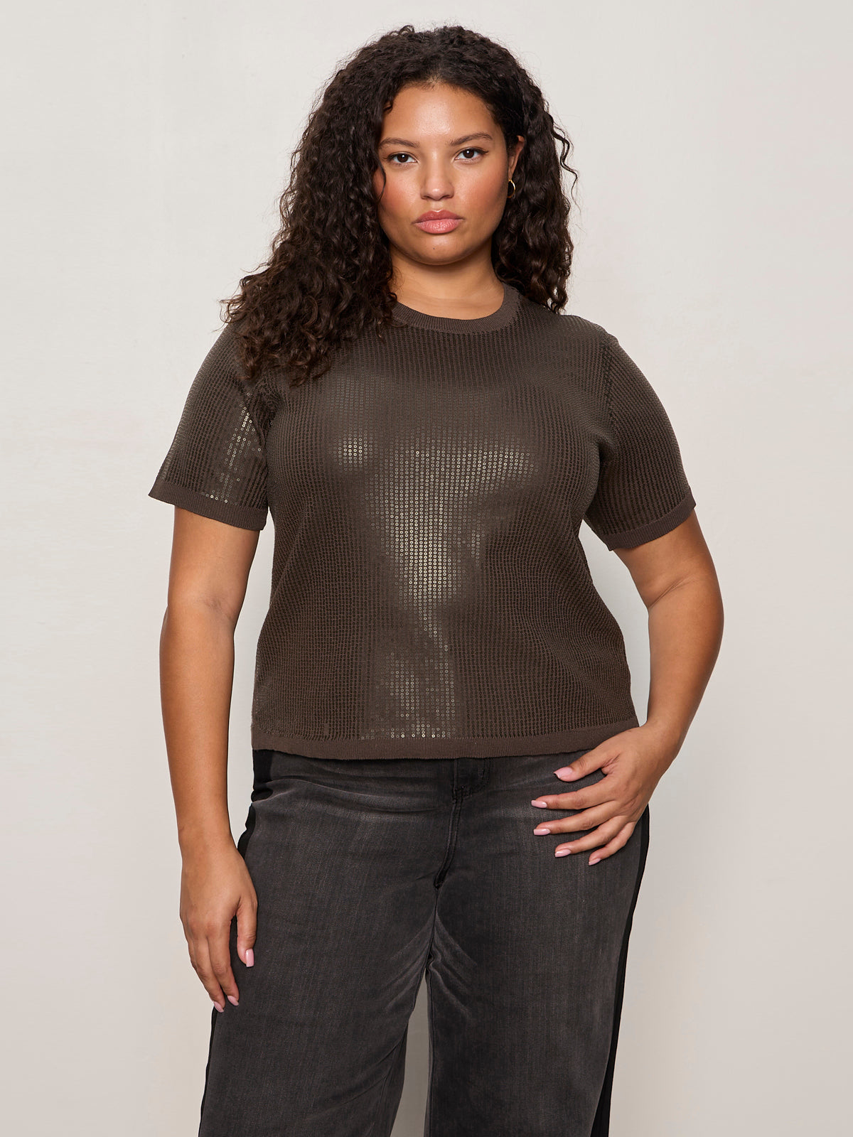 A woman with curly hair models the Sanctuary Clothing sparkle sequin sweater ink (extended sizing), paired with dark jeans, standing against a light-colored wall with one hand on her hip.
