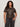 A woman with curly hair models the Sanctuary Clothing sparkle sequin sweater ink (extended sizing), paired with dark jeans, standing against a light-colored wall with one hand on her hip.