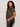 A woman with curly hair poses in profile against a plain light background, wearing the Sanctuary Clothing sparkle sequin sweater ink (extended sizing) with dark jeans, and maintains a neutral expression.
