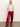 A woman stands against a plain background, wearing the Sanctuary Clothing sparkle sequin sweater mother of pearl, shiny red wide-leg pants with white side stripes, and red heeled boots. Her hands are in her pockets.
